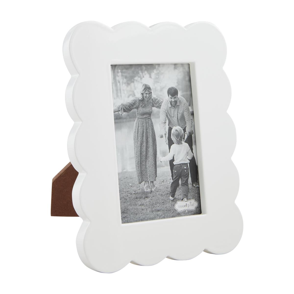 White scalloped photo frame with a brown easel back, displaying a black and white family portrait of a smiling mother, father, and toddler walking near a lake, featuring a small mud pie brand tag