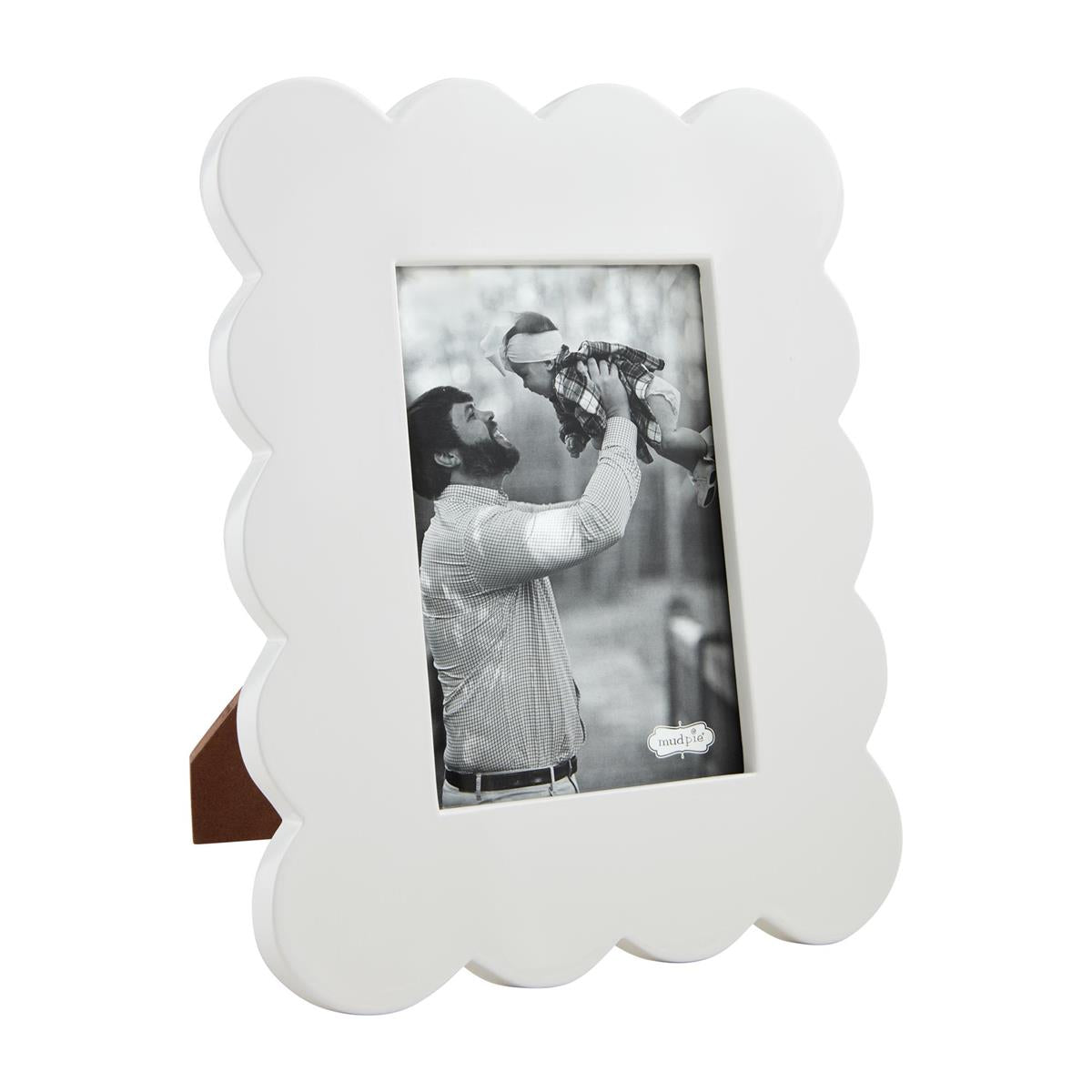 White mudpie scalloped photo frame with a black and white photo of a bearded father in a plaid shirt joyfully lifting a baby in a plaid outfit outdoors