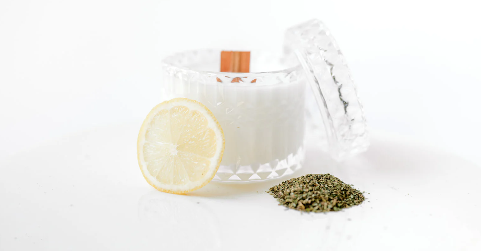 Glass candle jar with wooden wick next to a lemon slice and a small pile of dried herbs