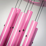 A close-up view of a modern pink wind chime featuring bright metallic fuchsia tubes suspended by thin black cords with small black grommets against a minimalist light background