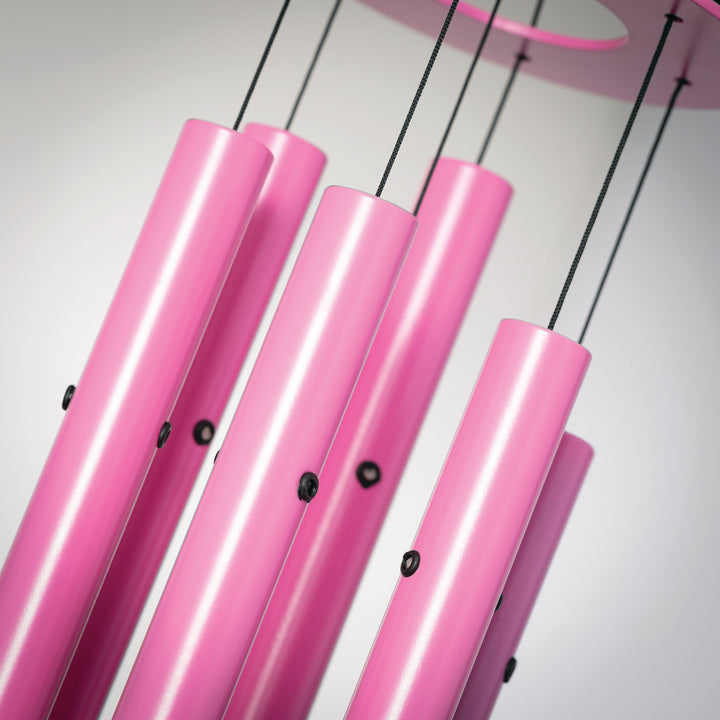 A close-up view of a modern pink wind chime featuring bright metallic fuchsia tubes suspended by thin black cords with small black grommets against a minimalist light background
