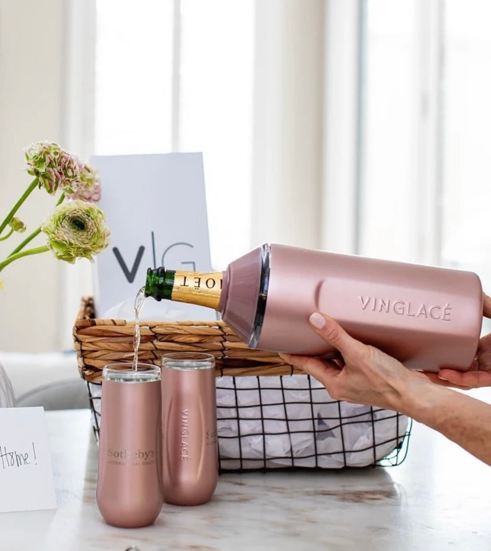 Hands pour Moët & Chandon champagne from a rose gold Vinglacé bottle chiller into two matching Sotheby's branded tumblers on a marble counter alongside a woven gift basket with green ranunculus flowers and a Home card