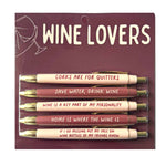 A packaged set of five "Wine Lovers" ballpoint pens, featuring cream and maroon barrels with gold tips, each inscribed with a humorous wine-themed quote such as "Corks are for quitters" or "Save water, drink wine", presented on a dark red cardboard backing with a white outline of a hand holding a wine glass