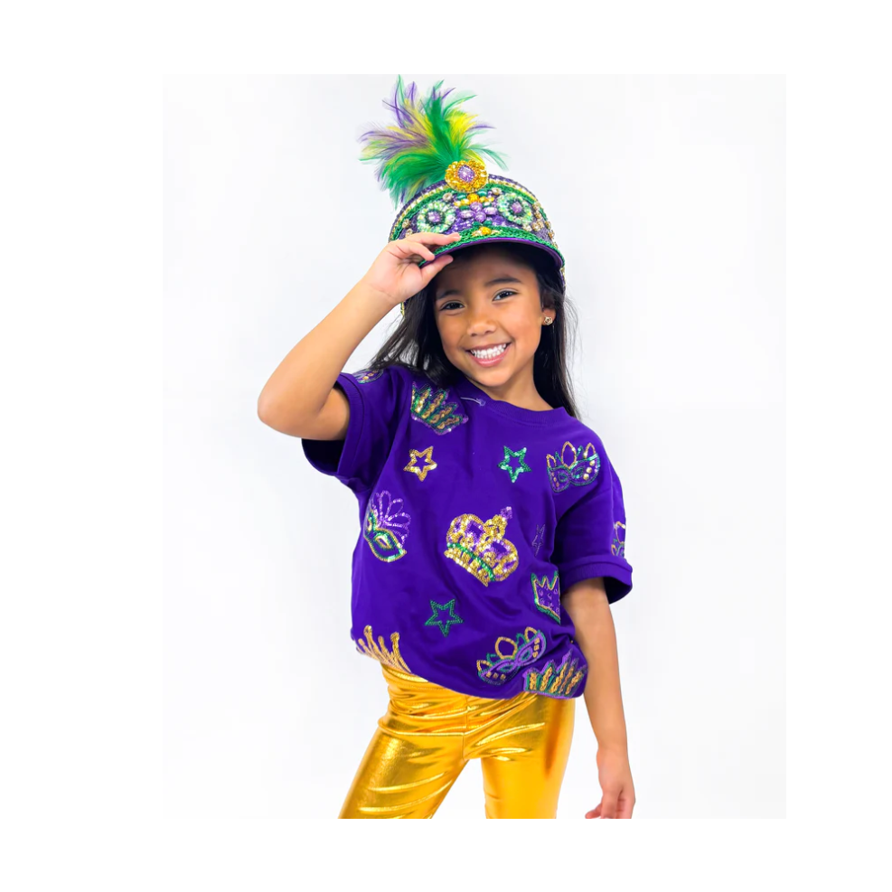 A smiling young girl in a studio dressed in a vibrant Mardi Gras outfit, featuring a purple, green, and gold feathered hat, a purple sequined t-shirt with masks and crowns, and shiny gold metallic leggings