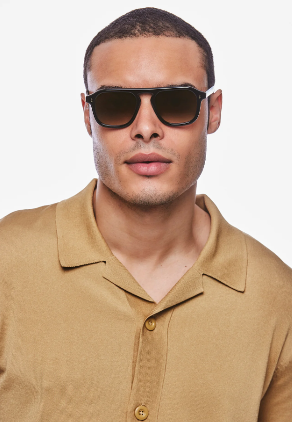 A man with dark skin wearing dark square-frame sunglasses with a double bridge and a tan knit polo shirt with an open collar on a white background