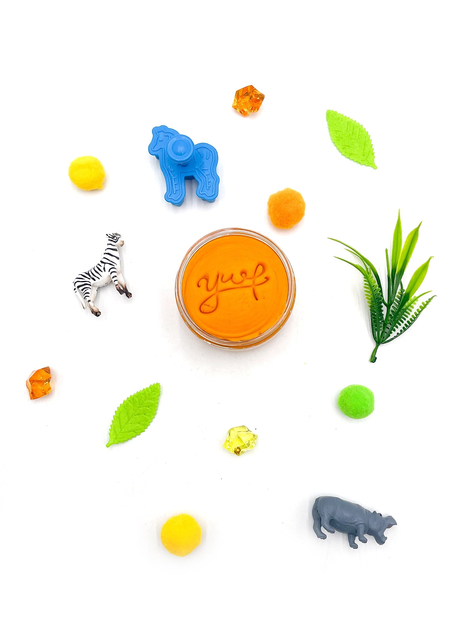 Flat lay of various children's safari-themed play items on a white background featuring an orange playdough jar with "yusf" on top, plastic zebra and hippo figures, a blue horse cookie cutter, and scattered green leaves, colorful pom-poms and crystal beads