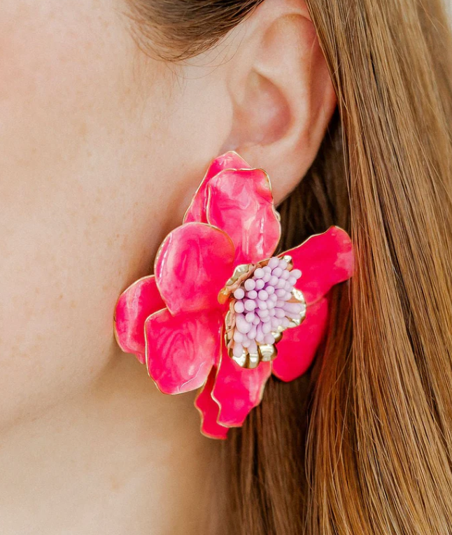 A hot pink glossy flower earring with a light purple beaded center and gold accents worn on a person's ear with light brown hair