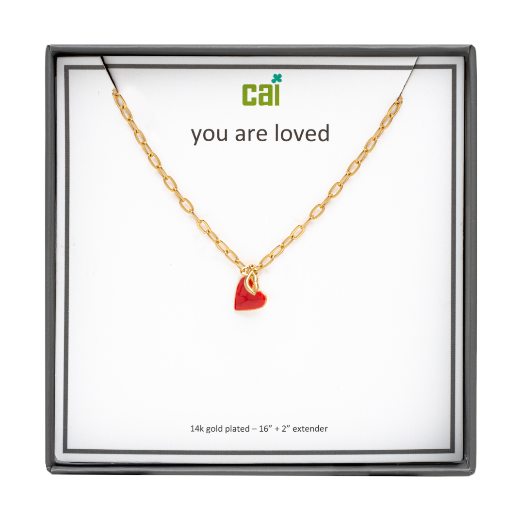 A gold-plated necklace with a red heart and gold lips pendant, presented in a display case with the text 'You are loved'.