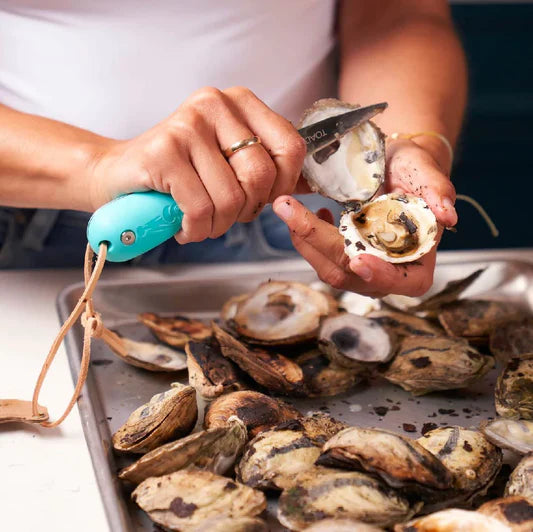 Person using a teal oyster knife to shuck an oyster over a metal tray filled with oysters
