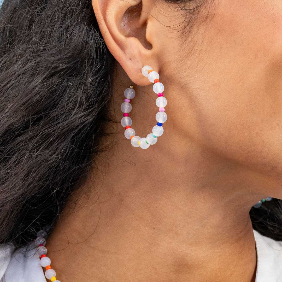 Close-up of a person's ear wearing a translucent white beaded hoop earring accented with small colorful beads in pink, red, orange, green, and blue with a matching necklace partially visible