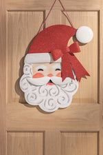 Decorative holiday sign of a smiling Santa Claus hanging on a wooden door, featuring a sparkling red glitter hat with a matching bow and a white beard accented with silver glitter swirls