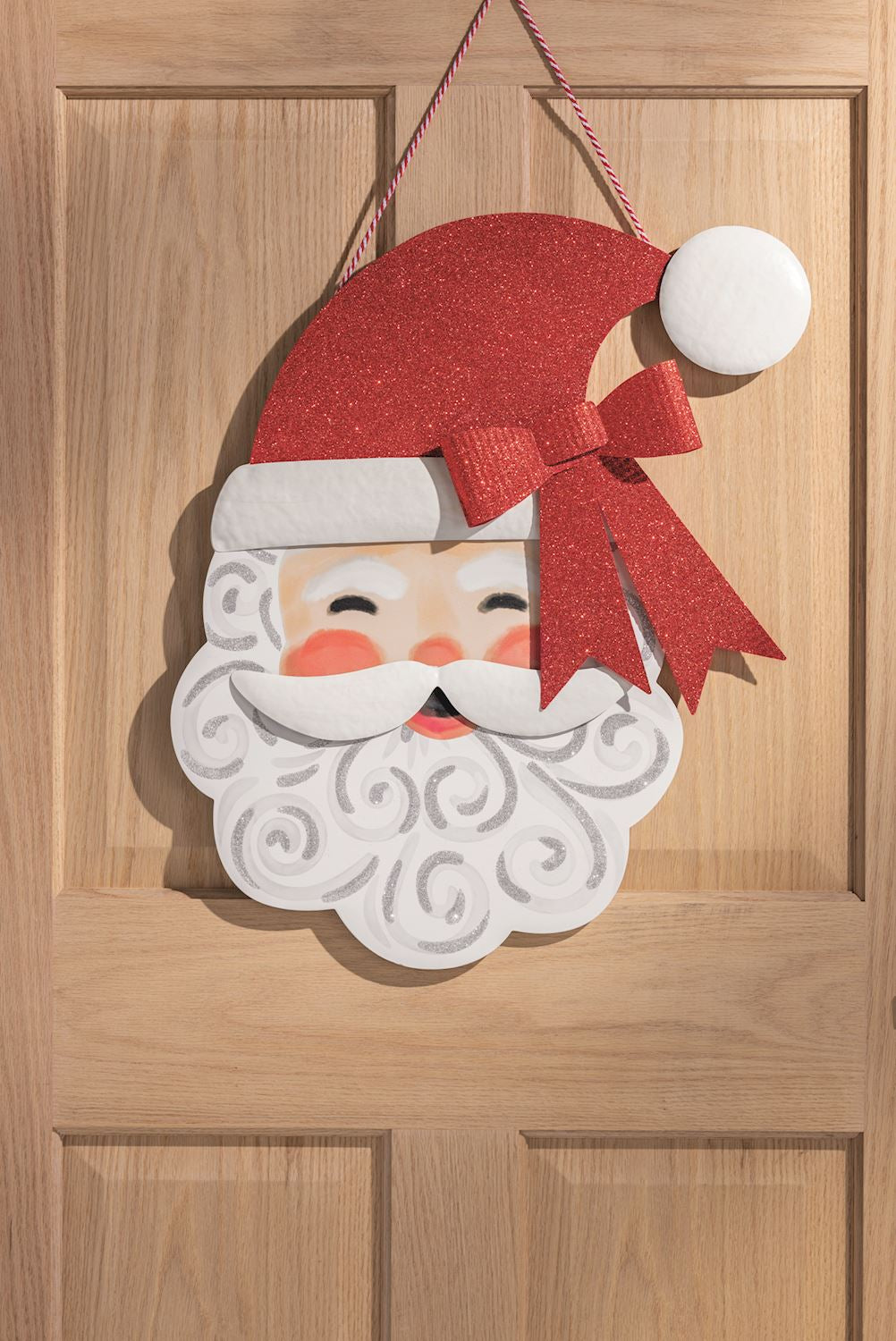 Decorative holiday sign of a smiling Santa Claus hanging on a wooden door, featuring a sparkling red glitter hat with a matching bow and a white beard accented with silver glitter swirls