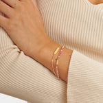 Two gold bracelets on a wrist one with a rectangular charm and one with pink beads