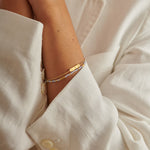Two layered gold bracelets with a small rectangular charm and light blue beads on a wrist