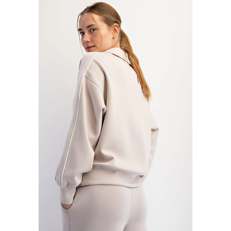 Young woman with a high ponytail and multiple ear piercings wearing a beige tracksuit with white sleeve stripes looking over her left shoulder in a plain studio setting her hand in a pocket