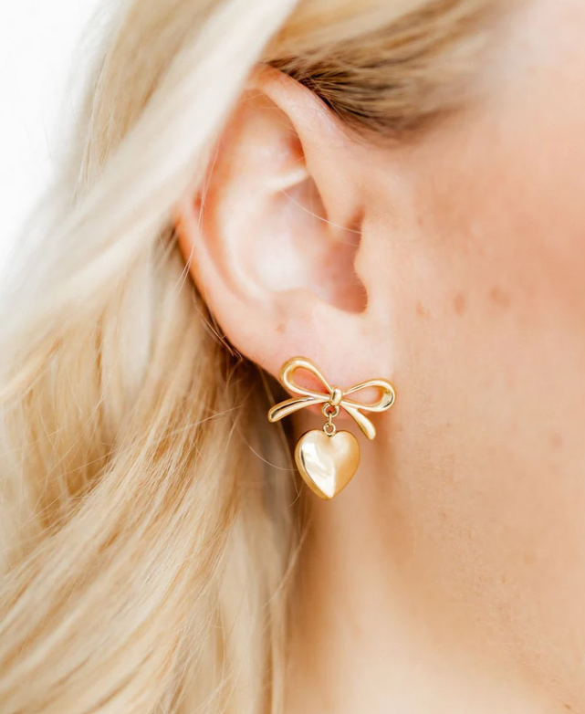 A shiny gold bow stud earring with a puffed heart dangle on a woman's ear with blonde hair