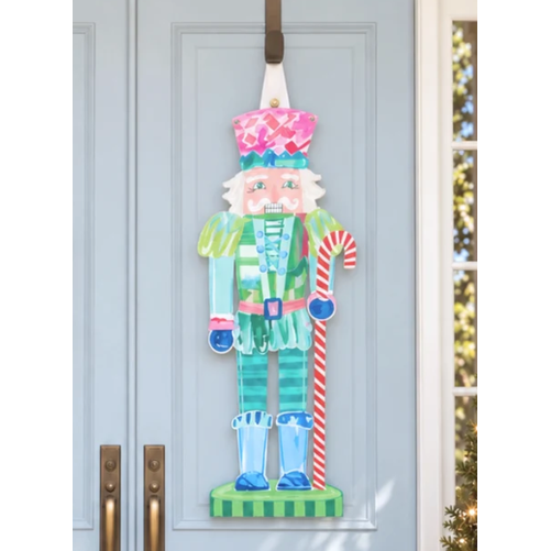 Painted nutcracker door hanger in pink, teal and green holding a candy cane on a light blue door