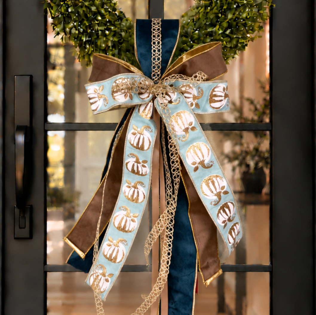 Close up of a decorative bow with pumpkin print and blue and brown ribbons attached to a green wreath