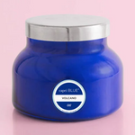 A blue candle labeled 'VOLCANO' by Capri Blue, placed on a pink surface.