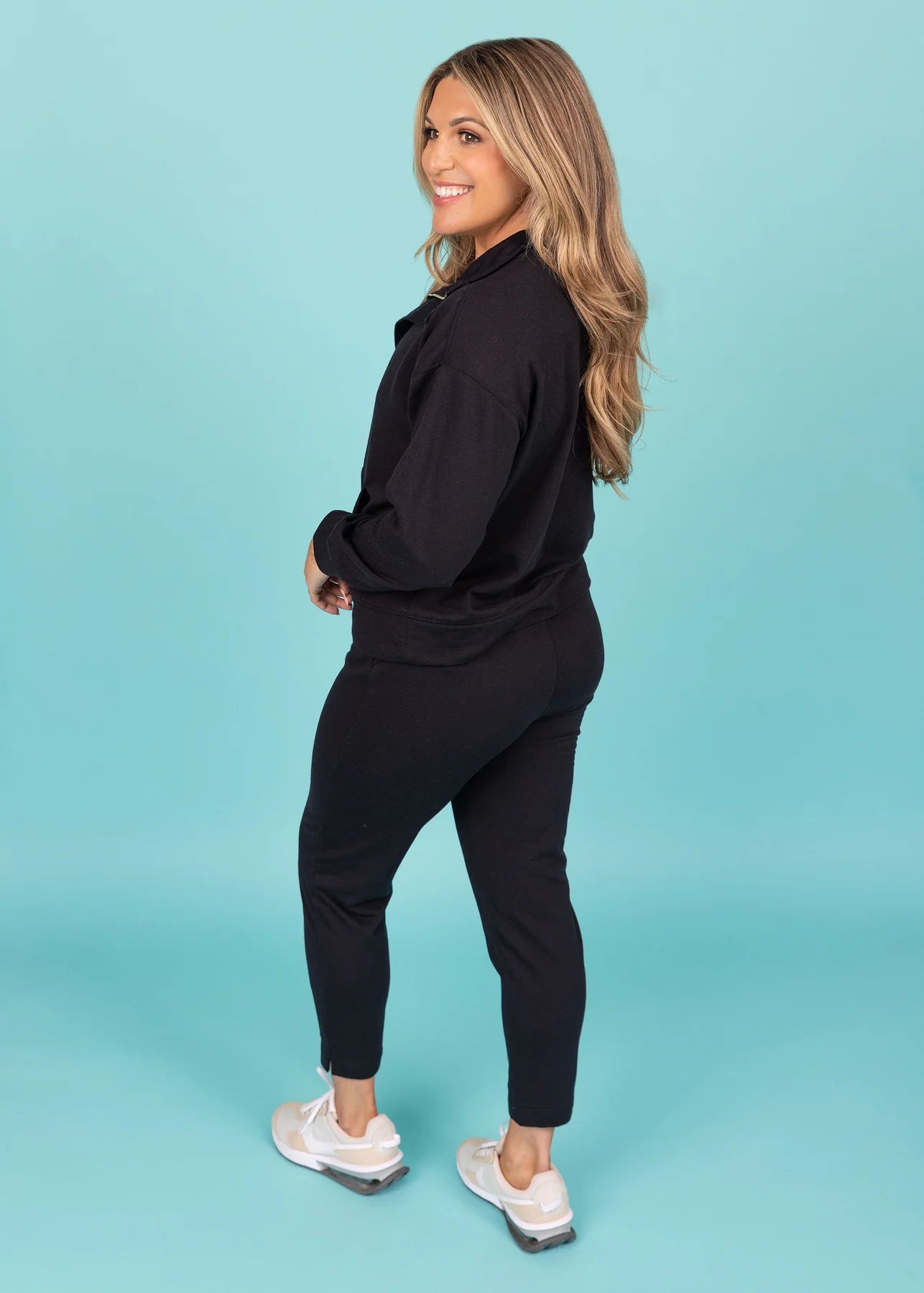 Photograph of a smiling woman with long blonde hair wearing a black loungewear set and light beige sneakers, turning over her shoulder against a vibrant light blue studio background