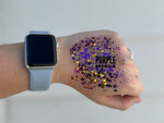Photograph of a hand wearing a silver Apple Watch with a light blue band, covered in a mix of purple, gold, and black hexagonal and dot glitter, with the text PURPLE AND GOLD overlaid