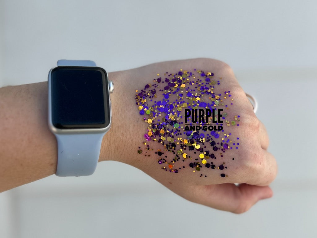 Photograph of a hand wearing a silver Apple Watch with a light blue band, covered in a mix of purple, gold, and black hexagonal and dot glitter, with the text PURPLE AND GOLD overlaid