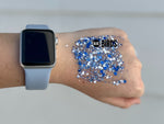 A close-up shot of a hand wearing a silver Apple Watch with a light blue band, next to a swatch of shimmering multi-sized blue and white chunky BLUE BIRDS glitter