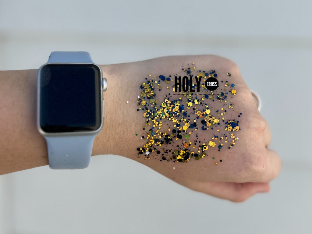 A hand displaying a light gray Apple Watch on the wrist and a patch of blue and gold glitter with a HOLY CROSS text overlay on the back
