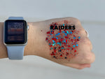 Close-up of a hand adorned with red, blue, and black hexagonal glitter and the faint text RAIDERS, along with an Apple Watch featuring a light blue band displaying 10:50 on the wrist