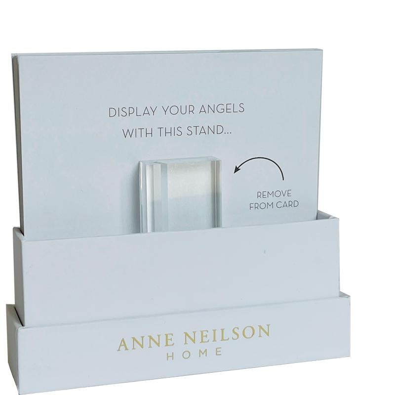 Anne Neilson Home product photography of a white cardboard display stand featuring a clear acrylic block, gold "ANNE NEILSON HOME" branding, and card text "DISPLAY YOUR ANGELS WITH THIS STAND" including a "REMOVE FROM CARD" instruction