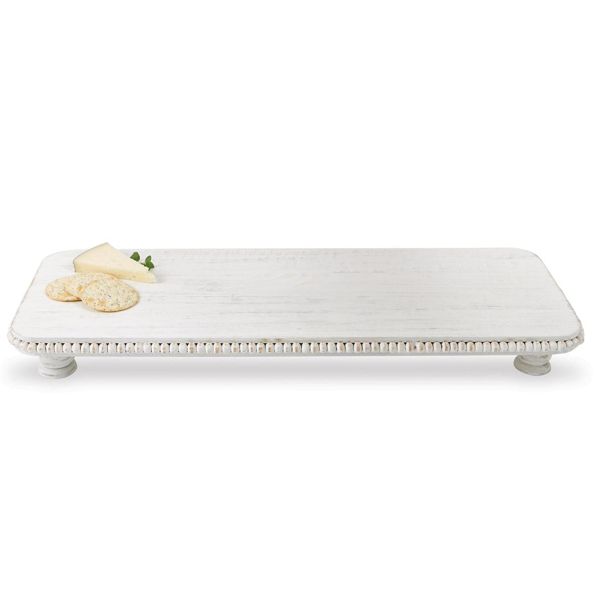 A rectangular distressed white wooden pedestal serving board featuring a carved beaded edge, supported by small round feet, showcasing a triangular cheese slice, three round crackers, and a fresh mint sprig on its surface