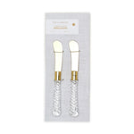 Product photography by Two's Company featuring a set of two crystal clear spreaders with gleaming gold metal blades and elegant twisted clear handles displayed on white branded packaging