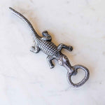 Silver-grey metallic lizard bottle opener featuring a textured scaly body resting on a white marble surface