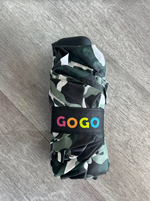 A folded compact umbrella with a camouflage pattern in green, black, and white rests on a gray wooden floor featuring a black strap with the colorful "GOGO" logo in pink, orange, yellow, and blue lettering