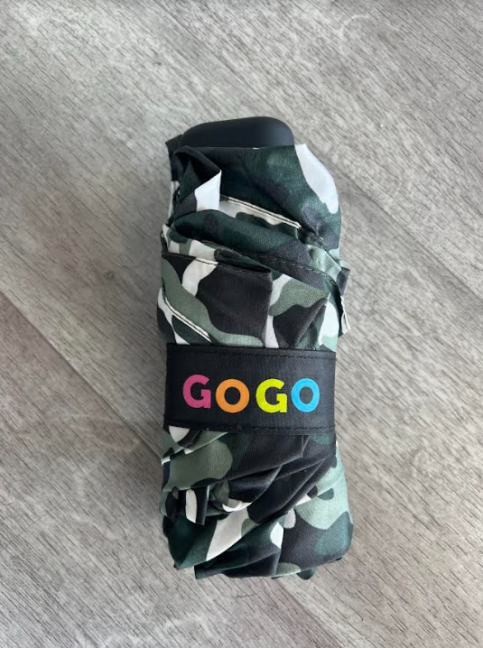 A folded compact umbrella with a camouflage pattern in green, black, and white rests on a gray wooden floor featuring a black strap with the colorful "GOGO" logo in pink, orange, yellow, and blue lettering