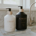 L'AVANT COLLECTIVE white hand lotion and black hand soap bottles with gold pumps and Fresh Linen branding displayed on a clear acrylic tray on a bathroom counter with a brushed nickel faucet