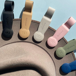 A studio shot featuring a semi-circular display of Klipsa magnetic hat clips in black, beige, light gray, blush pink, olive green, and royal blue, each embossed with the K logo and brand name, resting on a beige hat brim against a light blue background