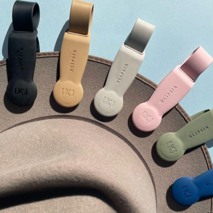 A studio shot featuring a semi-circular display of Klipsa magnetic hat clips in black, beige, light gray, blush pink, olive green, and royal blue, each embossed with the K logo and brand name, resting on a beige hat brim against a light blue background