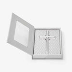 A polished silver cross resting inside an open white gift box featuring a clear window and a light gray pattern of interconnected crosses