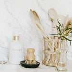 A kitchen counter featuring a white L'AVANT Collective dish soap bottle with gold pump a wooden scrub brush on a black soap dish a rattan holder filled with wooden spoons and a clear glass vase with a green eucalyptus branch set against a white marble backsplash