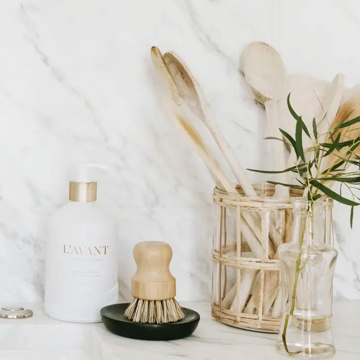 A kitchen counter featuring a white L'AVANT Collective dish soap bottle with gold pump a wooden scrub brush on a black soap dish a rattan holder filled with wooden spoons and a clear glass vase with a green eucalyptus branch set against a white marble backsplash