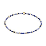 Jewelry bracelet featuring small round gold beads alternating with short cylindrical dark blue and light blue lavender beads, centered by a single larger gold bead on a clean white background