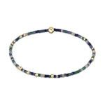 Photograph of a delicate beaded bracelet composed of tiny rectangular beads in shades of navy blue, light blue, dark green, and pale grey, interspersed with small and a central prominent shiny gold spherical beads on a white background