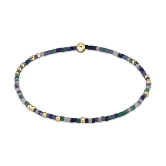 Photograph of a delicate beaded bracelet composed of tiny rectangular beads in shades of navy blue, light blue, dark green, and pale grey, interspersed with small and a central prominent shiny gold spherical beads on a white background