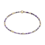 Beaded bracelet with small seed beads in shades of lavender light purple white and grey interspersed with tiny gold accent beads and a larger central gold sphere on a white background
