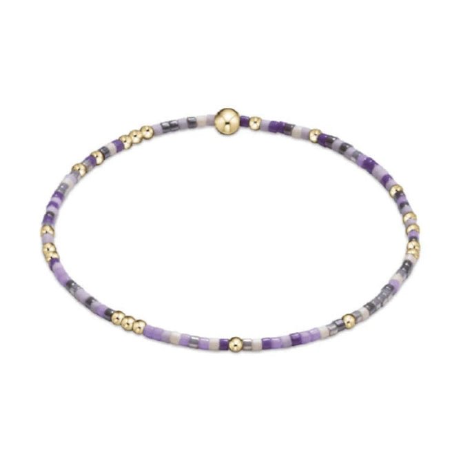 Beaded bracelet with small seed beads in shades of lavender light purple white and grey interspersed with tiny gold accent beads and a larger central gold sphere on a white background