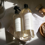 Archipelago plant-based laundry spot cleaner and detergent bottles on a folded white terry cloth towel, accompanied by a wooden scrubbing brush and a woven basket with wooden clothespins, all on a bright white surface bathed in strong sunlight with sharp shadows