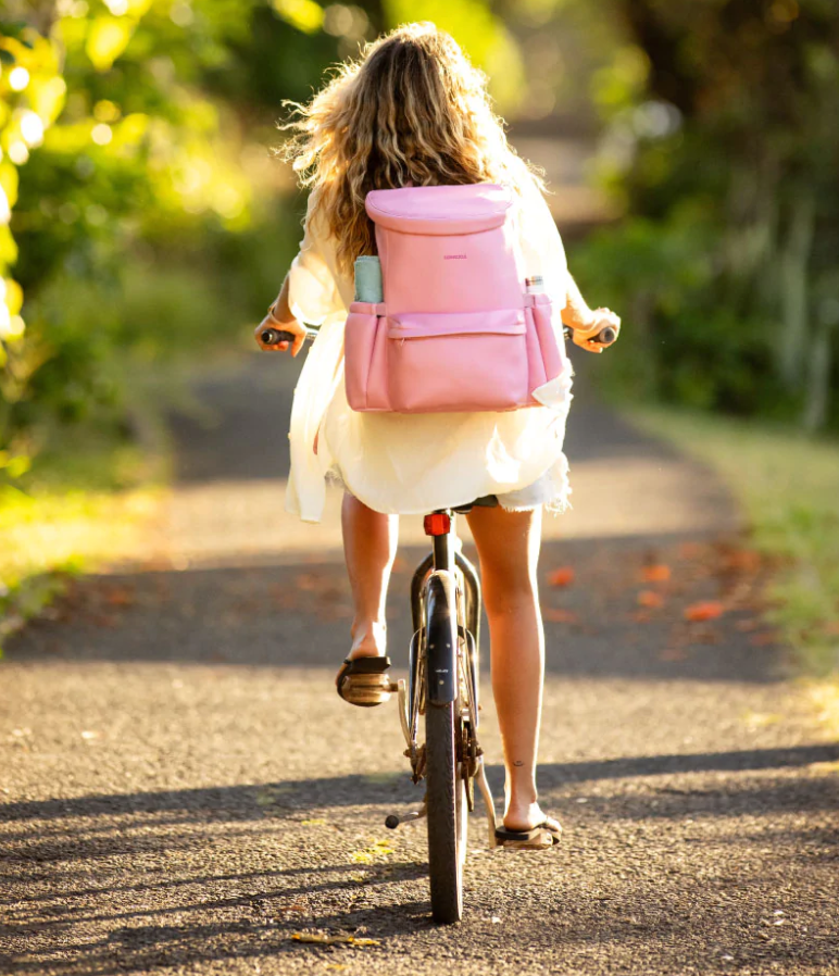 Photography of a woman with wavy blonde hair riding a bicycle on a sunlit path, wearing a flowy white top and a vibrant pink Konkola backpack holding a mint green bottle