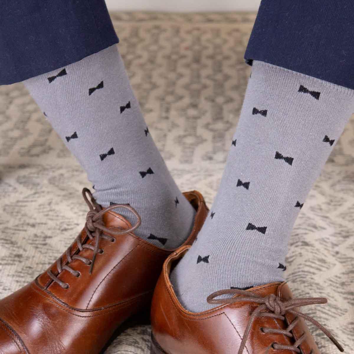Photograph of a person's legs wearing light gray socks with black bow tie patterns, shiny brown leather oxford shoes, and navy blue trousers on a neutral patterned rug