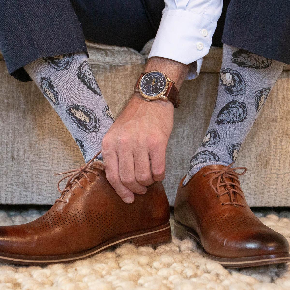 A person fastening a brown leather oxford shoe while wearing light grey socks with black and tan oyster patterns, dark blue trousers, a white shirt, and a brown leather watch, standing on a cream rug